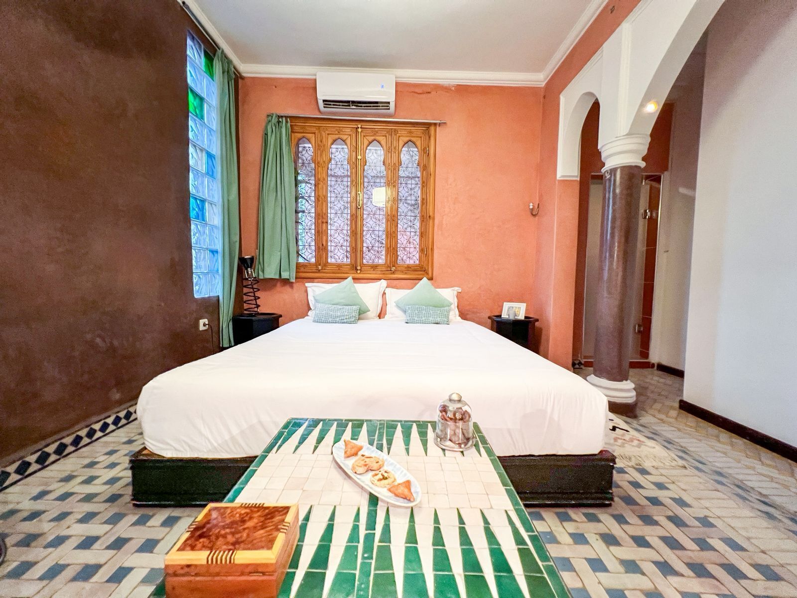 A hotel room with a large bed, a small table with food, a window with curtains, and a tiled floor.