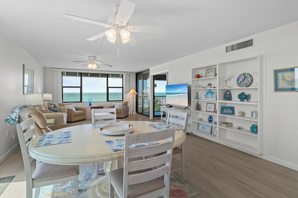 Un salon spacieux avec une grande table à manger, des sièges confortables, un ventilateur de plafond et une vue sur l’océan grâce à de grandes fenêtres.
