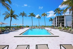 A pool area with lounge chairs and umbrellas, palm trees, and a building in the background.