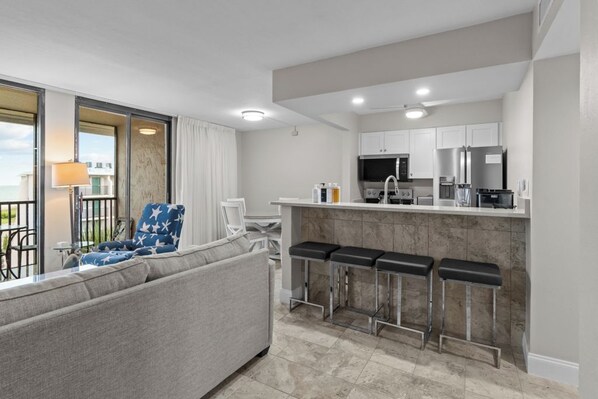Una habitación de hotel moderna con sofá, una zona de bar con taburetes, una cocina con armarios blancos y vistas al mar a través de un ventanal grande.