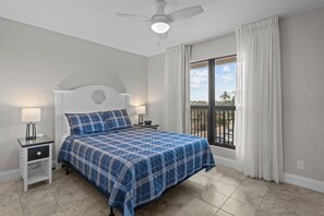 A bedroom with a bed, nightstands, a ceiling fan, a window with curtains, and a view of the outdoors.