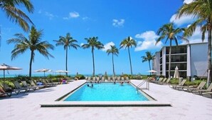 A pool area with lounge chairs and umbrellas, palm trees, and a building in the background.