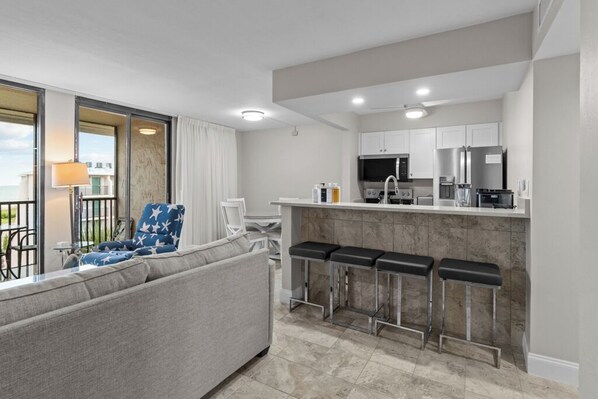 Una habitación de hotel moderna con sofá, una zona de bar con taburetes, una cocina con armarios blancos y vistas al mar a través de un ventanal grande.