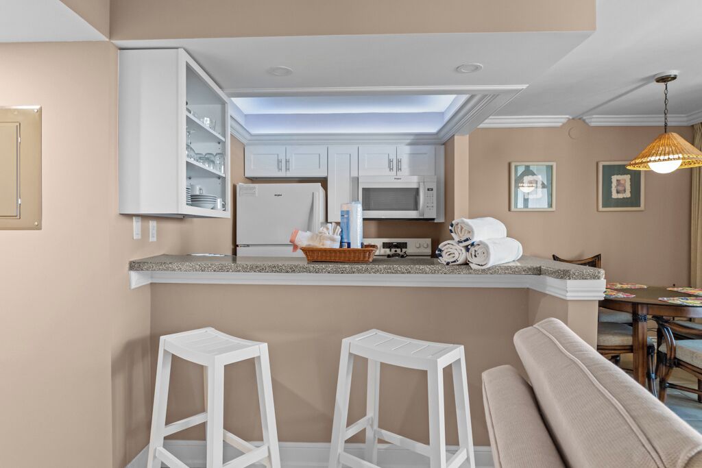 Een moderne keuken met witte kasten, een magnetron en een koelkast. Op de voorgrond staan twee witte barstoelen en een bankje.