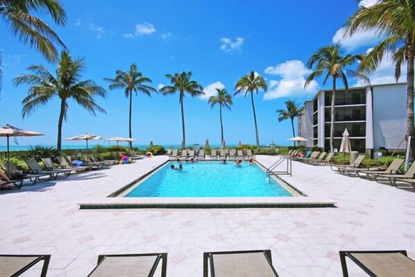 A pool area with lounge chairs and umbrellas, palm trees, and a building in the background.