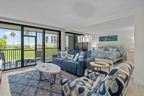 A modern living room with a blue sofa, a round coffee table, and a patterned armchair. There is a bed with a blue and white bedspread and a painting above it. The room has large glass doors leading to a balcony with a view of palm trees and a building.