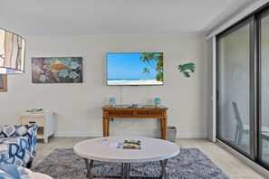 A living room with a round table, a sofa, a TV on the wall, and a view of the beach through a sliding door.