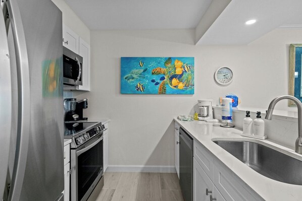 A modern kitchen with stainless steel appliances, a microwave, a coffee maker, and a sink. There is a painting of a sea turtle and fish on the wall.