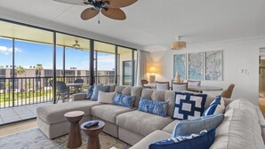 A spacious living room with a large sectional sofa, a dining area, and a balcony with a view of palm trees.
