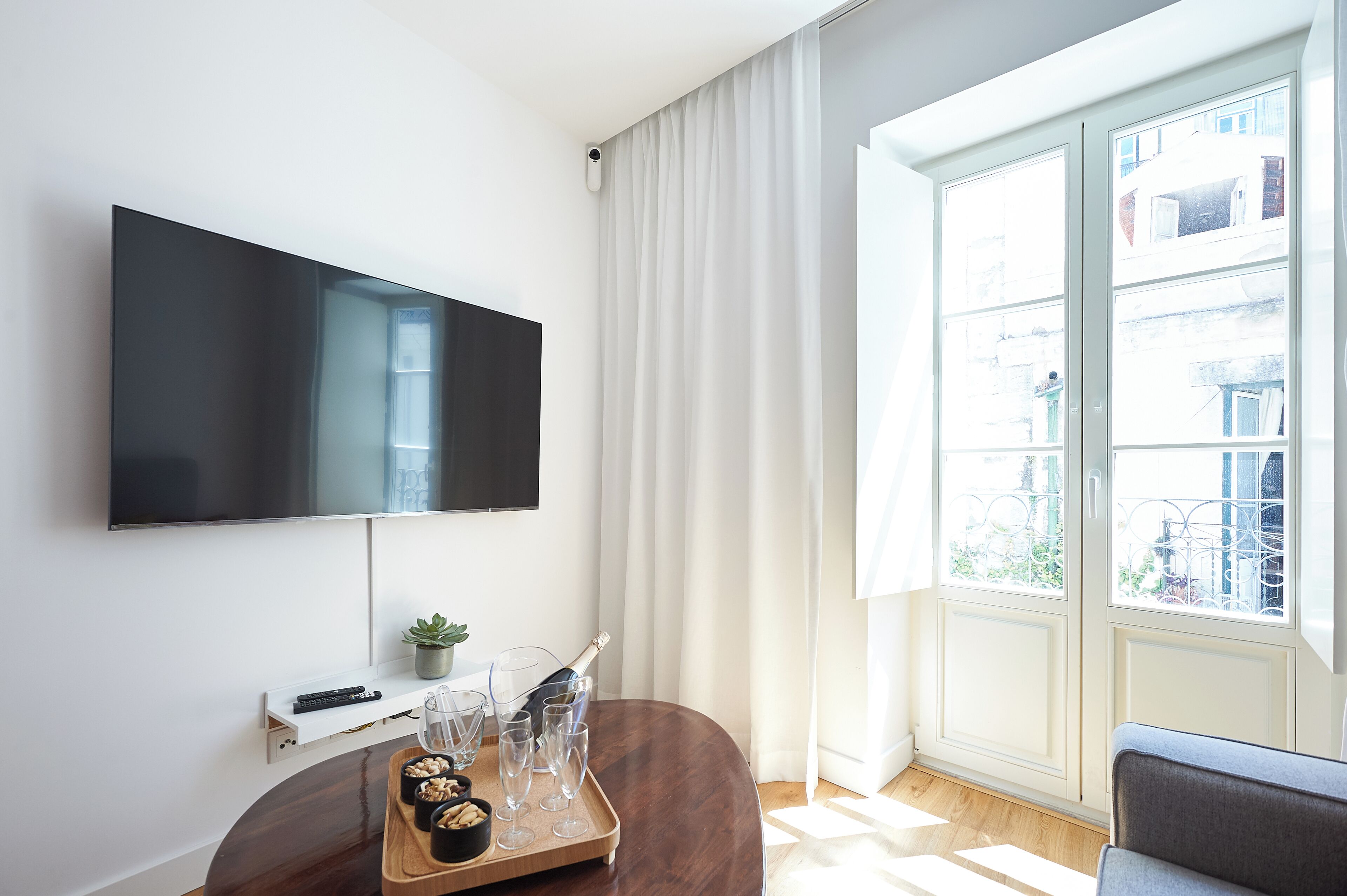 A modern living room with a flat-screen TV mounted on the wall, a wooden table with a tray of snacks and glasses, a small potted plant, and a large window with curtains.