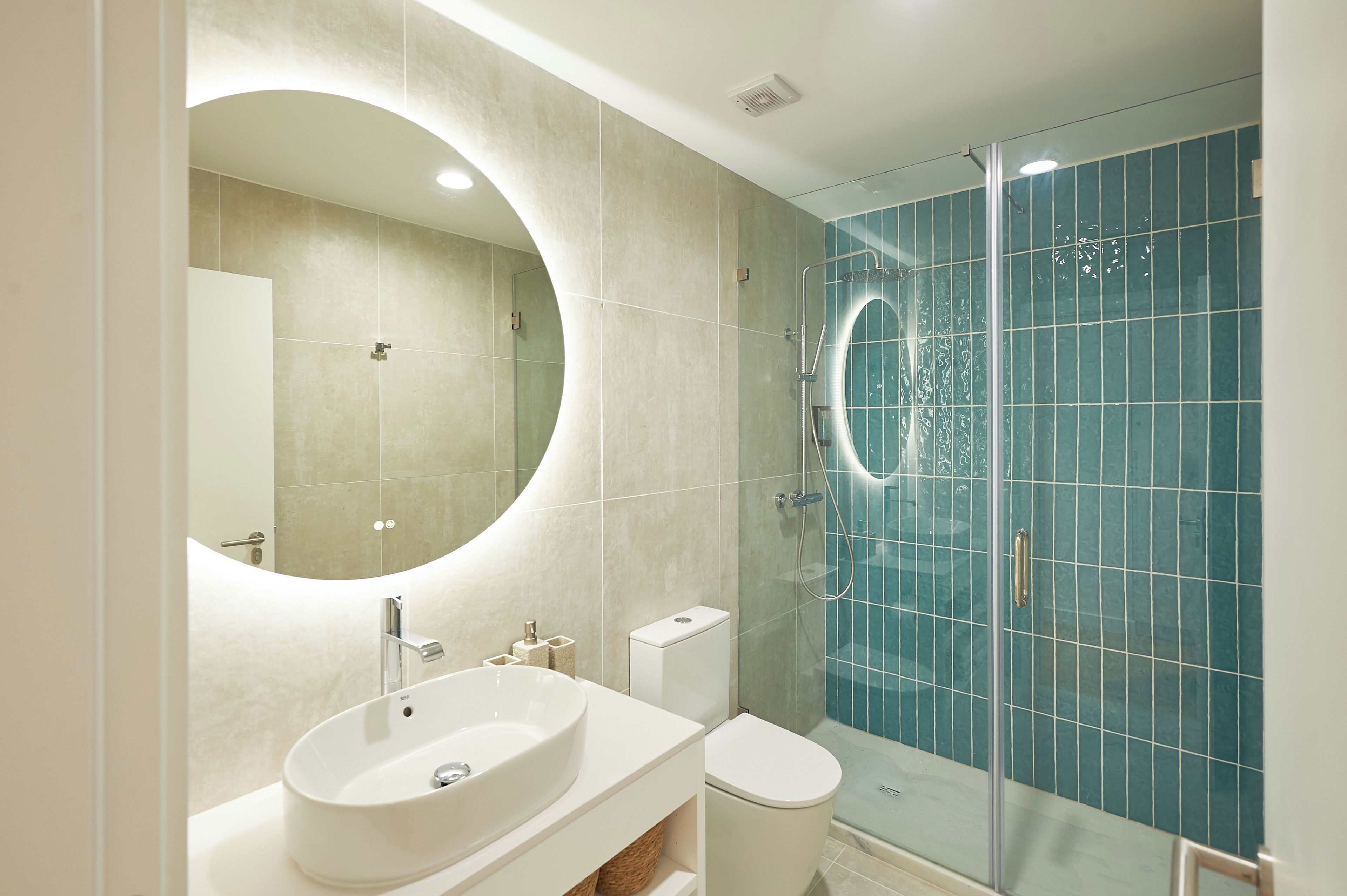 A modern bathroom with a large oval mirror, a white sink, a toilet, and a glass-enclosed shower area.