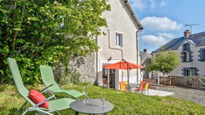 Un espace extérieur aménagé avec des chaises vertes et une table, un parasol rouge et un mur de pierre en arrière-plan.