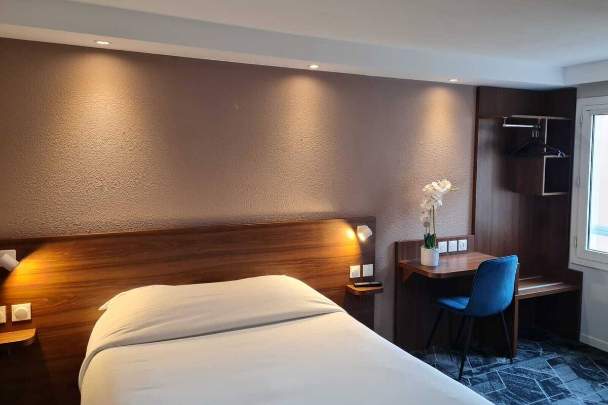 A hotel room with a large bed, a desk with a chair, a small table with a vase of flowers, and a wardrobe.