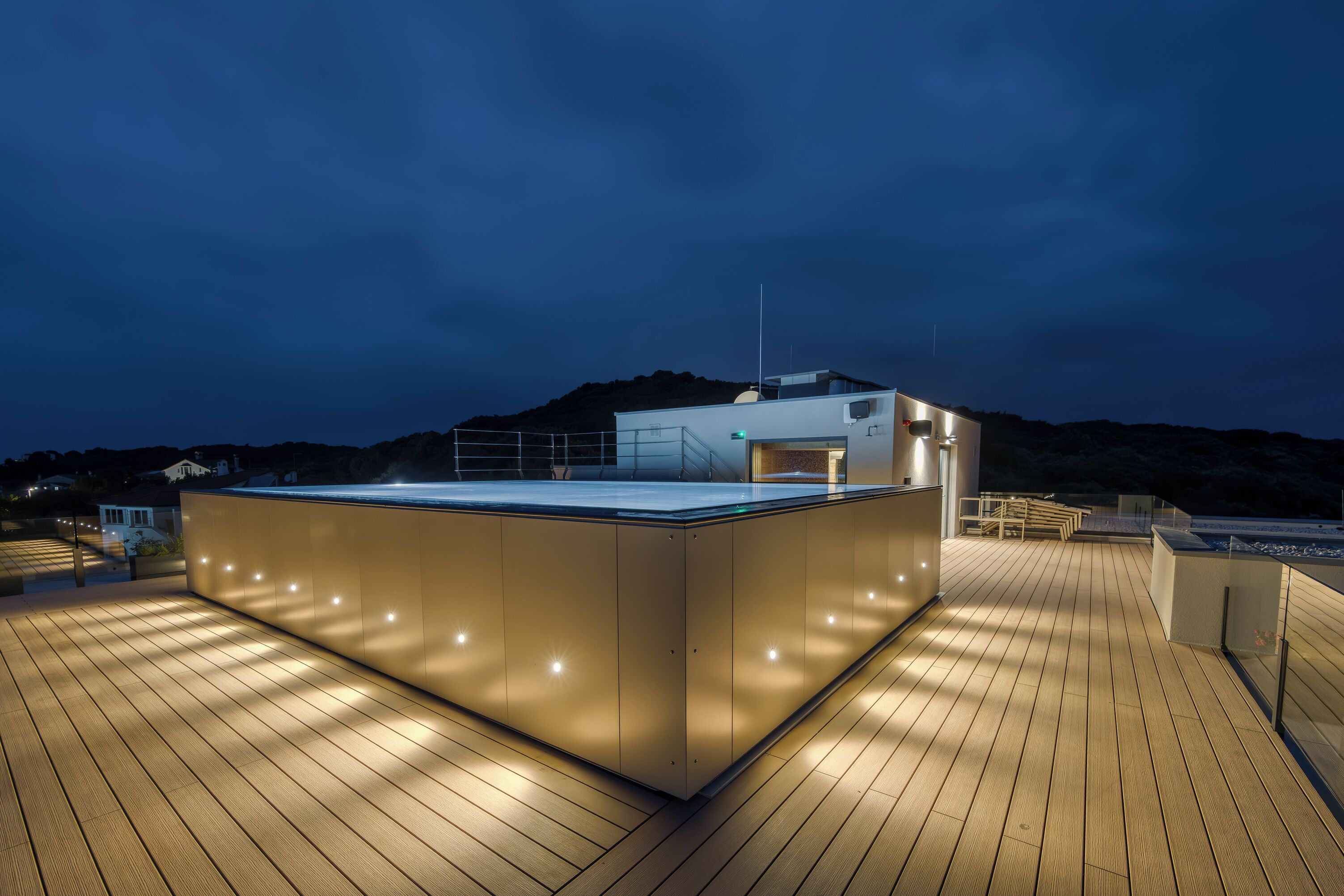 A modern rooftop deck with a wooden floor and a glass enclosure, surrounded by a railing and a mountain backdrop.
