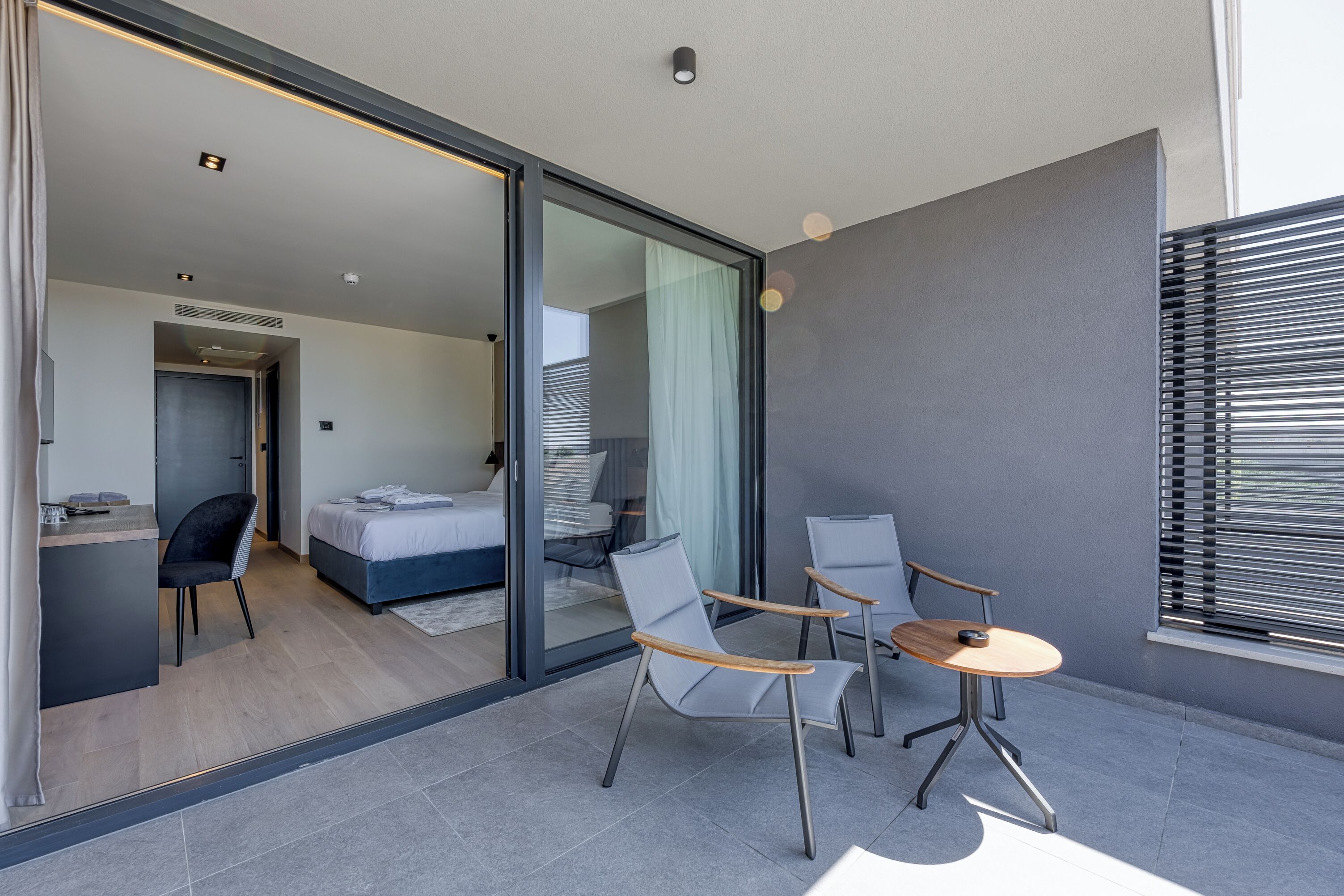 A modern hotel room with a glass door leading to a balcony. The room includes a bed, a desk, and two chairs.