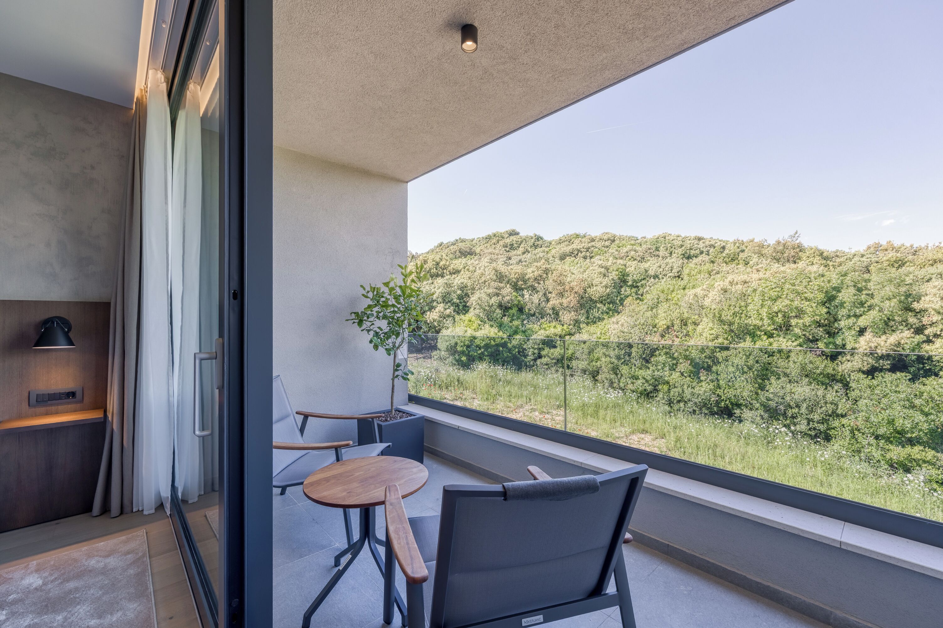 A balcony with a view of a forested hillside, a small table, and a chair.