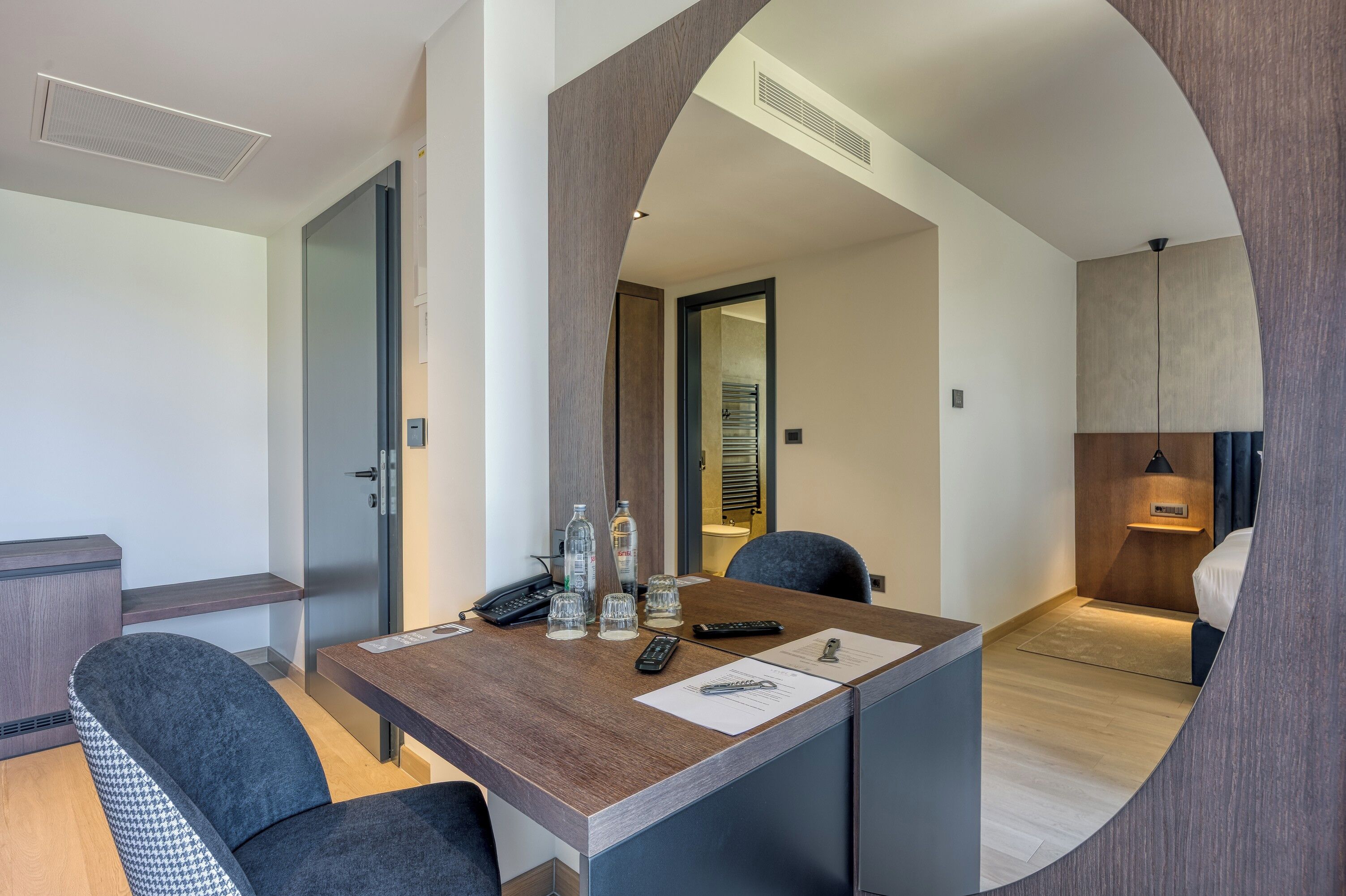 A modern hotel room with a wooden desk, two chairs, a minibar, and a bathroom visible through an archway.