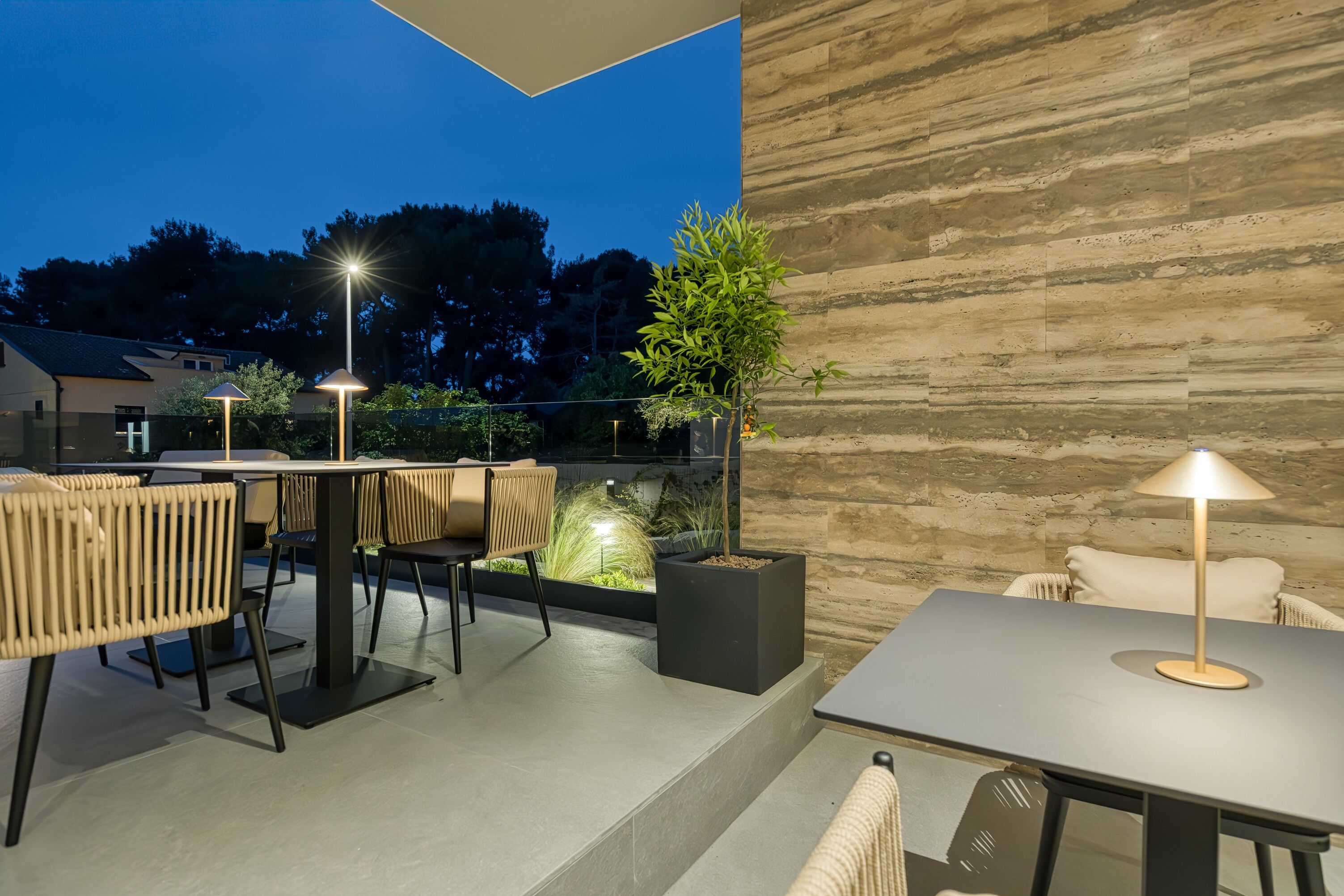 An outdoor patio area with tables and chairs, a potted plant, and a stone wall.