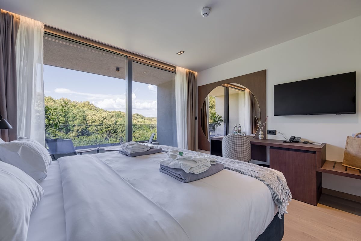 Ein modernes Hotelzimmer mit einem großen Bett, einem Schreibtisch mit Flachbildfernseher und Blick auf eine üppige, grüne Landschaft.