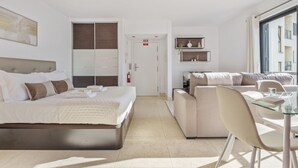 Ein modernes Hotelzimmer mit einem großen Bett, einer Schlafcouch, einem Glastisch und einer kleinen Küchenzeile.