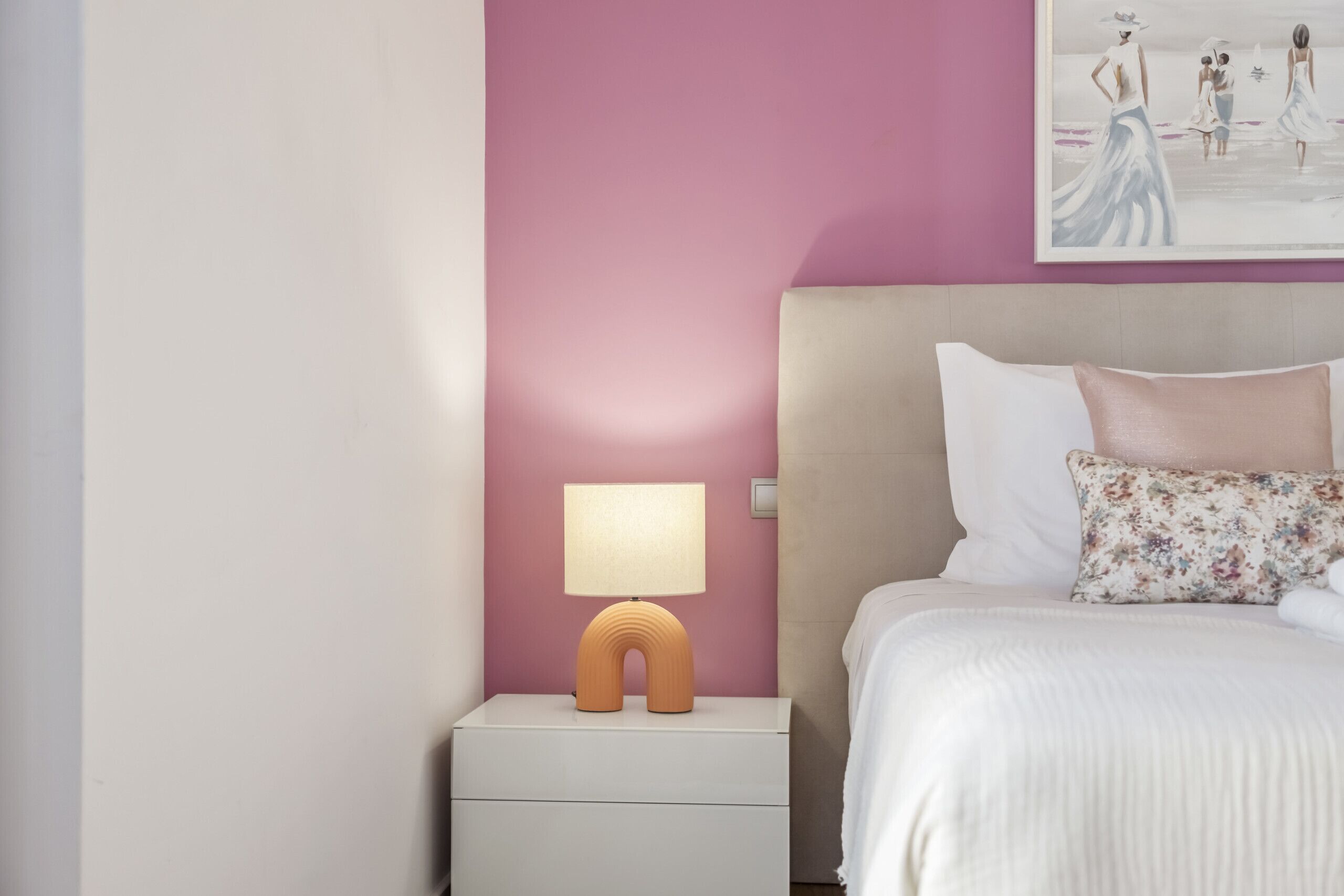A bedroom with a pink wall, a white nightstand with a unique lamp, a bed with white bedding and a floral pillow, and a painting above the bed.