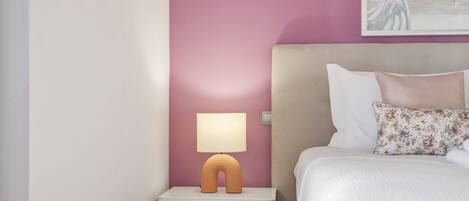 A bedroom with a pink wall, a white nightstand with a unique lamp, a bed with white bedding and a floral pillow, and a painting above the bed.