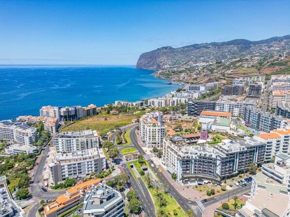 Un paysage urbain côtier avec des bâtiments modernes, une mer bleue et limpide, et un arrière-plan vallonné.