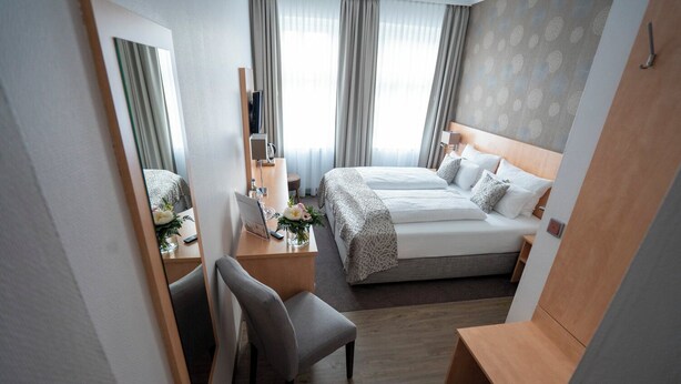 Una habitación de hotel con cama, escritorio con ordenador, silla, espejo y ventana con cortinas.