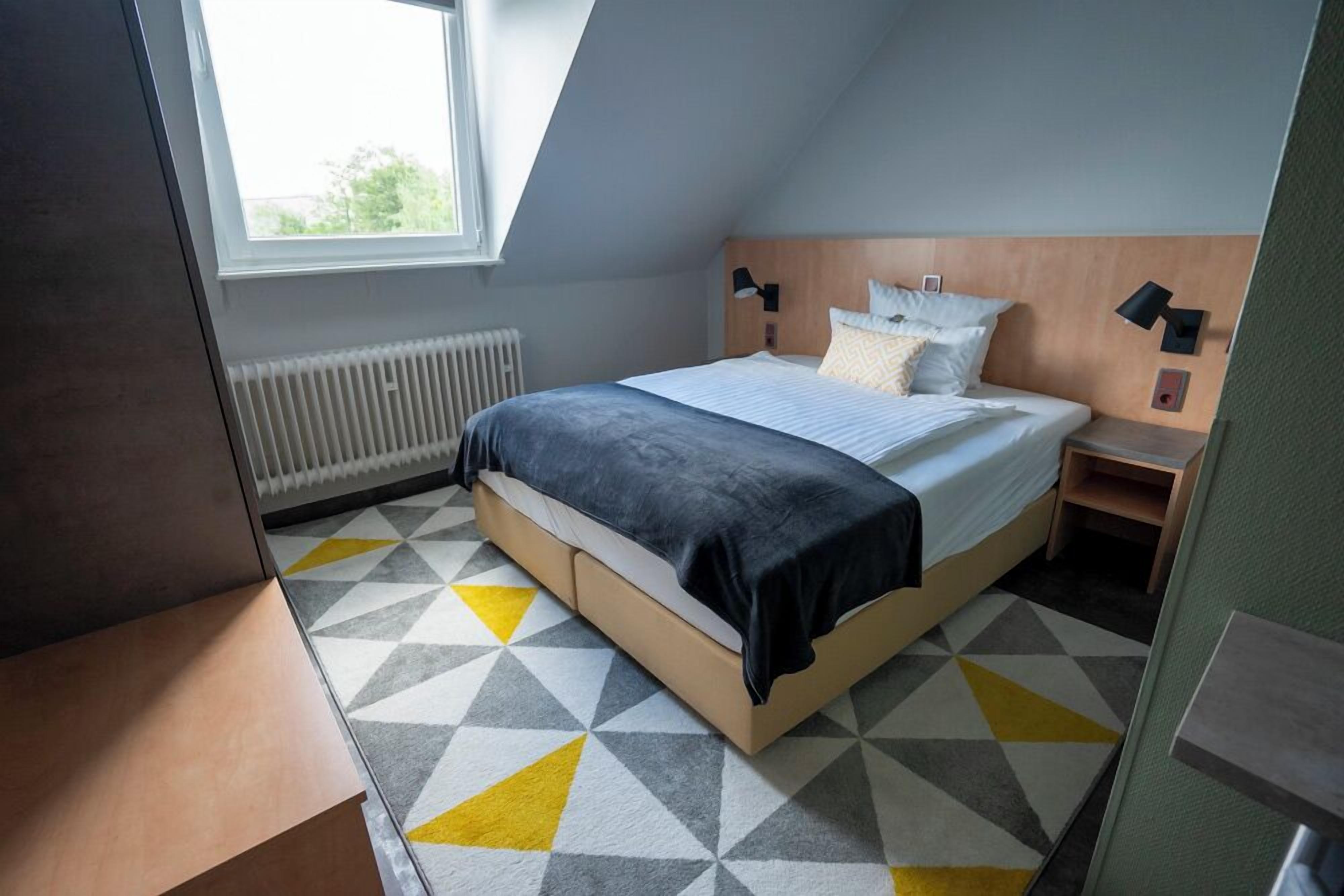 Quarto moderno com teto inclinado, uma cama grande com cabeceira de madeira, luminárias de cabeceira e um tapete com padrão geométrico.