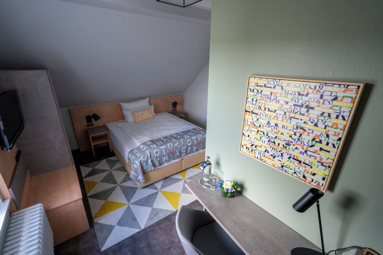 A modern hotel room with a bed, a desk, a chair, a TV, and a wall art piece.
