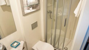 Un baño pequeño con inodoro, cabina de ducha de cristal, espejo y un estante con artículos de aseo.