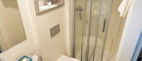 A compact bathroom with a toilet, a glass shower enclosure, a mirror, and a small shelf with toiletries.