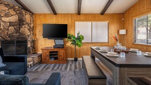 A cozy living room with a stone fireplace, a flat-screen TV, wooden walls, a dining area with a bench, and a large window. - Moonridge Villa, BBQ, Spa, Wood Fireplace (Big Bear)