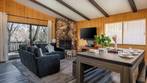 A cozy living room with a stone fireplace, a black sofa, and a dining table set for six. - Moonridge Villa, BBQ, Spa, Wood Fireplace (Big Bear)