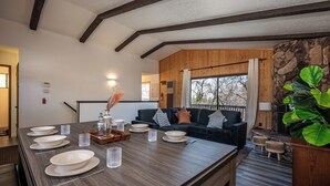 A modern dining area with a long wooden table set for six, a dark-colored sofa, and a large green plant. - Moonridge Villa, BBQ, Spa, Wood Fireplace (Big Bear)