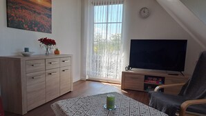 Uma sala de estar acolhedora com um armário de madeira, uma televisão, uma mesa com toalha de renda e uma janela com cortinas.
