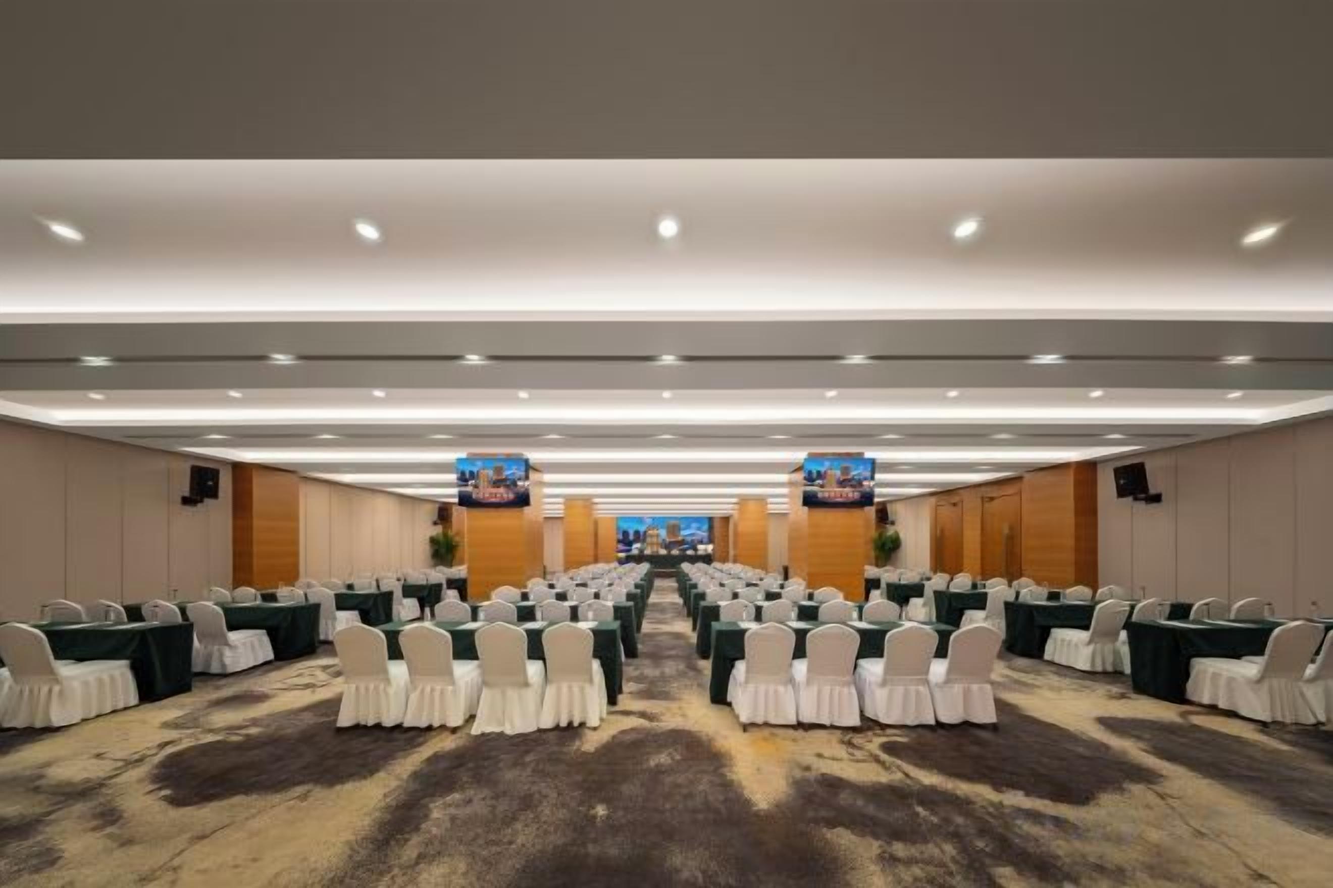 A conference room with rows of tables and chairs, a carpeted floor, and a high ceiling with recessed lighting.