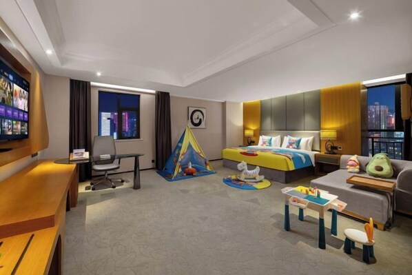 Habitación de hotel con cama, escritorio, televisión, ventana con cortinas, vista a la ciudad y un pequeño espacio de juegos.