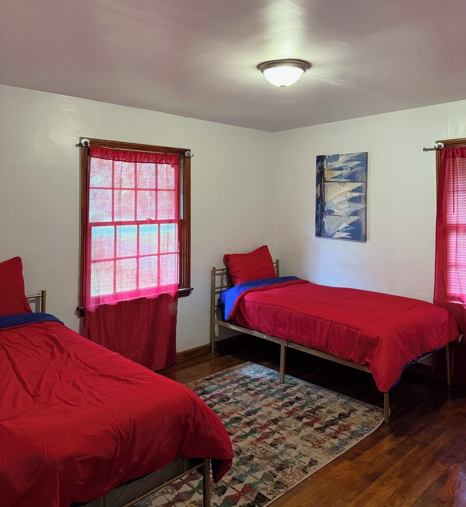 Une chambre avec deux lits simples, un plancher en bois, un tapis à motifs, un luminaire au plafond et un tableau accroché au mur.