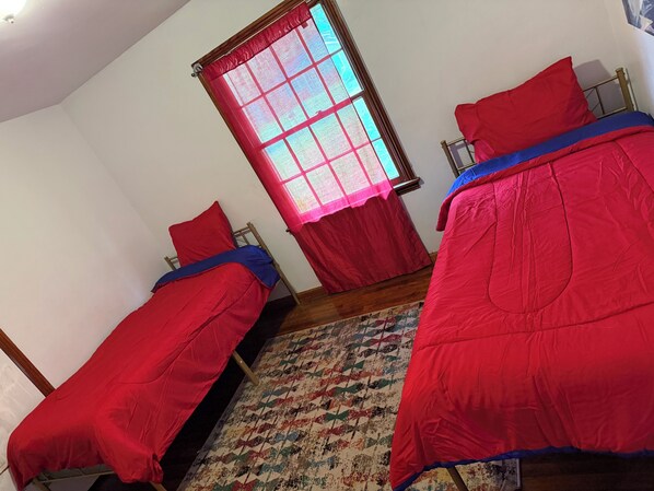 Une chambre avec deux lits simples, chacun étant équipé de draps rouges et d’une couverture bleue. Il y a une fenêtre avec un rideau rouge et un tapis à motifs sur le sol.