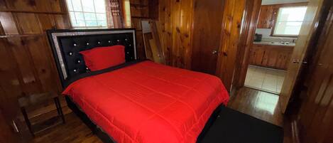 A bedroom with a wooden paneled wall, a bed with a red bedspread, a nightstand, and a wooden door.