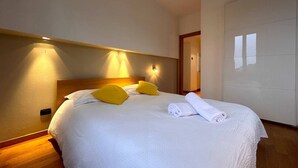 Un lit bien fait, avec une literie blanche et deux coussins jaune vif, une tête de lit en bois et un luminaire fixé au mur.