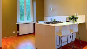 Une petite cuisine avec une fenêtre, un radiateur et un plan de travail blanc, ainsi que deux chaises.