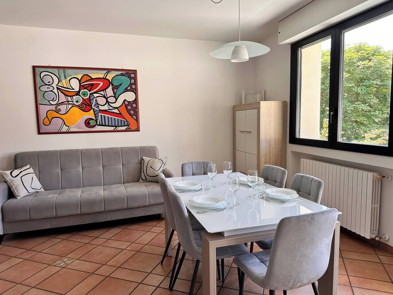 Un área de comedor con una mesa blanca dispuesta para cuatro personas, un sofá gris y un cuadro abstracto colorido en la pared.