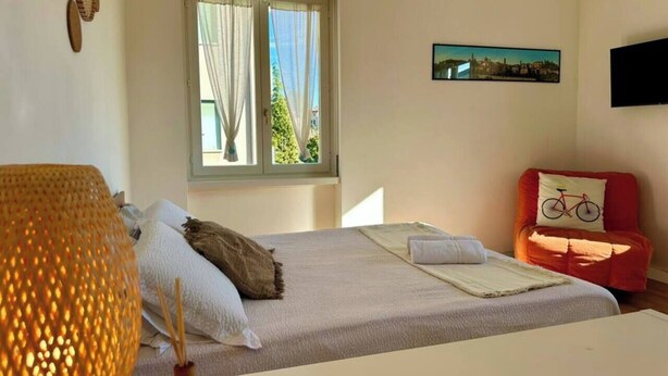 A bedroom with a bed, a window with curtains, a red chair with a bicycle pillow, and a wall-mounted TV.