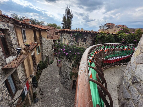 A narrow cobblestone street flanked by stone buildings with balconies, a wrought iron fence, and a flag.