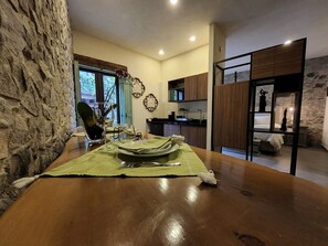 Un comedor con una mesa de madera, sillas y un mantel individual verde. Se observa una pared de piedra en un lado y una zona de cocina al fondo.