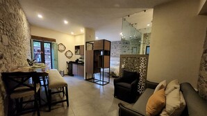 A modern living room with a sofa, a dining table with chairs, a kitchen area, and a bedroom with a glass door.
