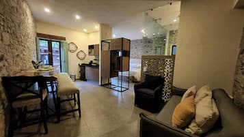 A modern living room with a sofa, a dining table with chairs, a kitchen area, and a bedroom with a glass door.