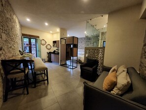 A modern living room with a sofa, a dining table with chairs, a kitchen area, and a bedroom with a glass door.